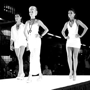 Models pose in white outfits on a fashion runway stage during a vintage style show against a dark background.