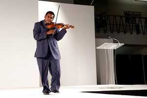 Violinist performs on stage wearing a navy blue suit against a white backdrop.  Dallas World Trade Center.