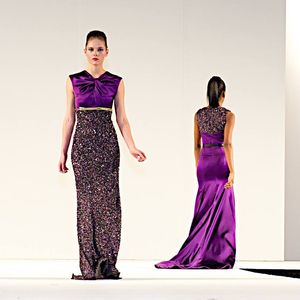 Models showcase elegant evening gowns with purple satin and sequined details on a white runway stage.