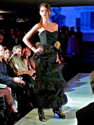 Model walks runway in black strapless evening gown with layered ruffles during fashion show event.