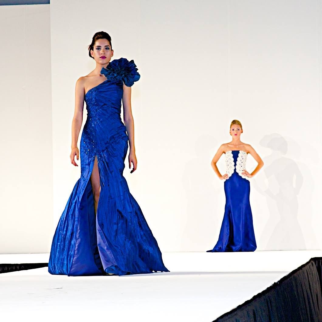 Model showcases elegant royal blue evening gown with dramatic one-shoulder ruffle detail and thigh-high slit on fashion runway.  WTC.