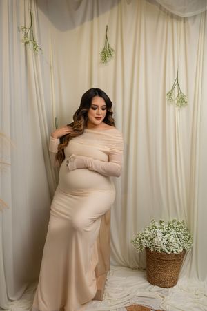 Elegant maternity photo featuring an off-shoulder cream gown against draped fabric with hanging greenery and wicker basket decor.