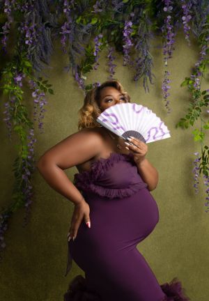 Elegant maternity portrait under hanging wisteria with purple dress and white lace fan against sage green backdrop.