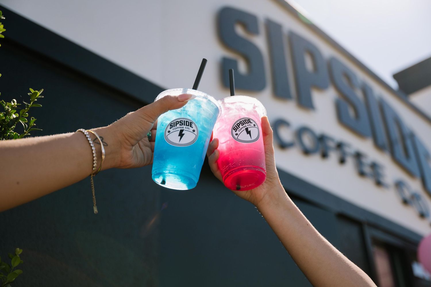 Two hands toasting colorful blue and pink iced drinks outside Sipside Coffee Co on a sunny day.