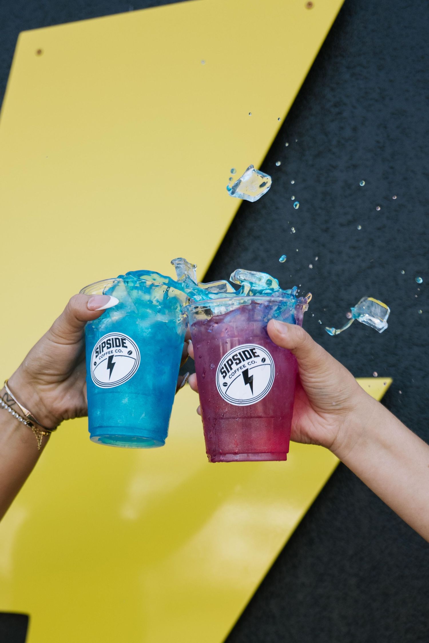 Two hands toasting colorful blue and pink Sipside drinks, splashing liquid against a yellow and black background.