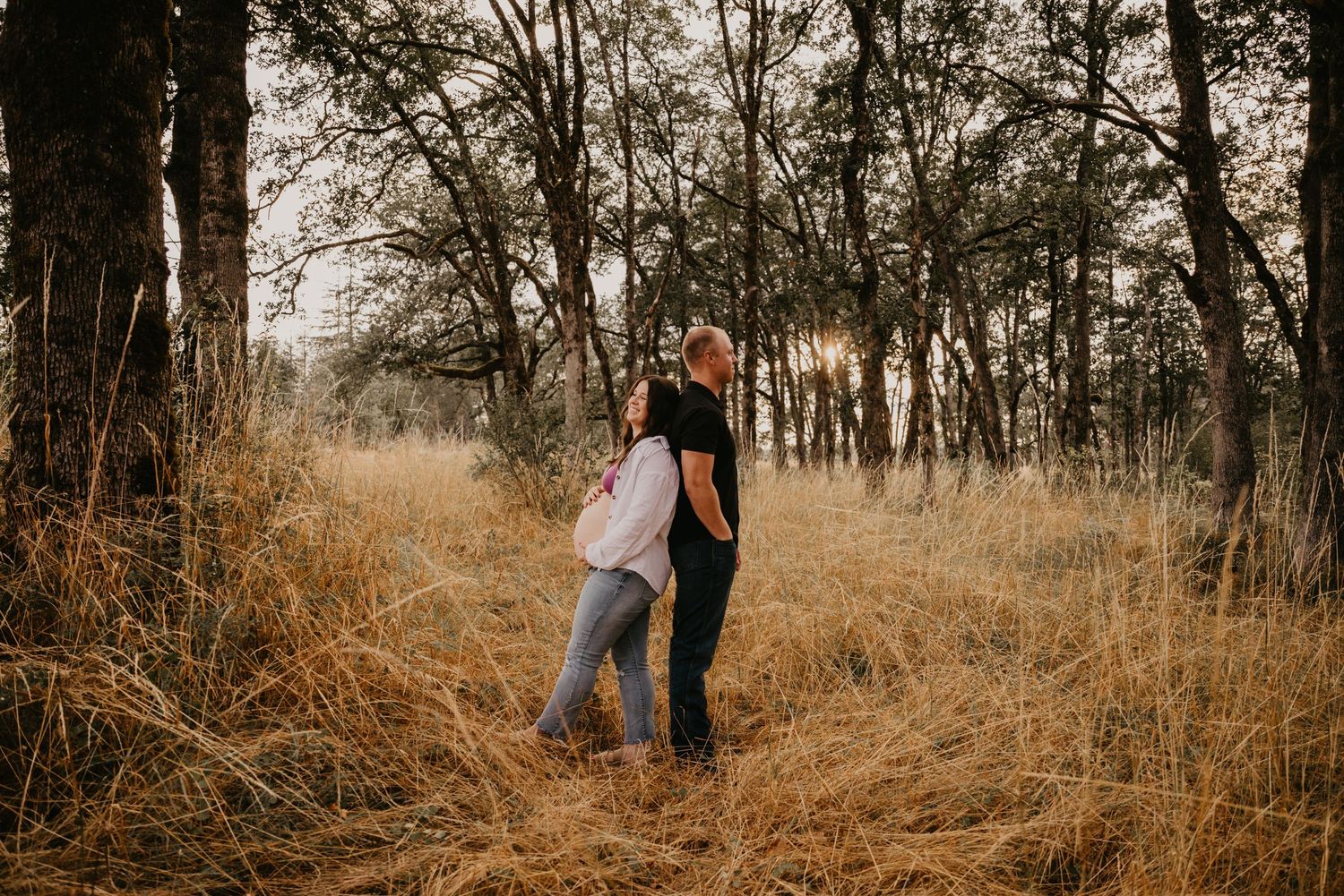 A romantic couple embraces in a field of tall golden grass surrounded by trees during sunset.