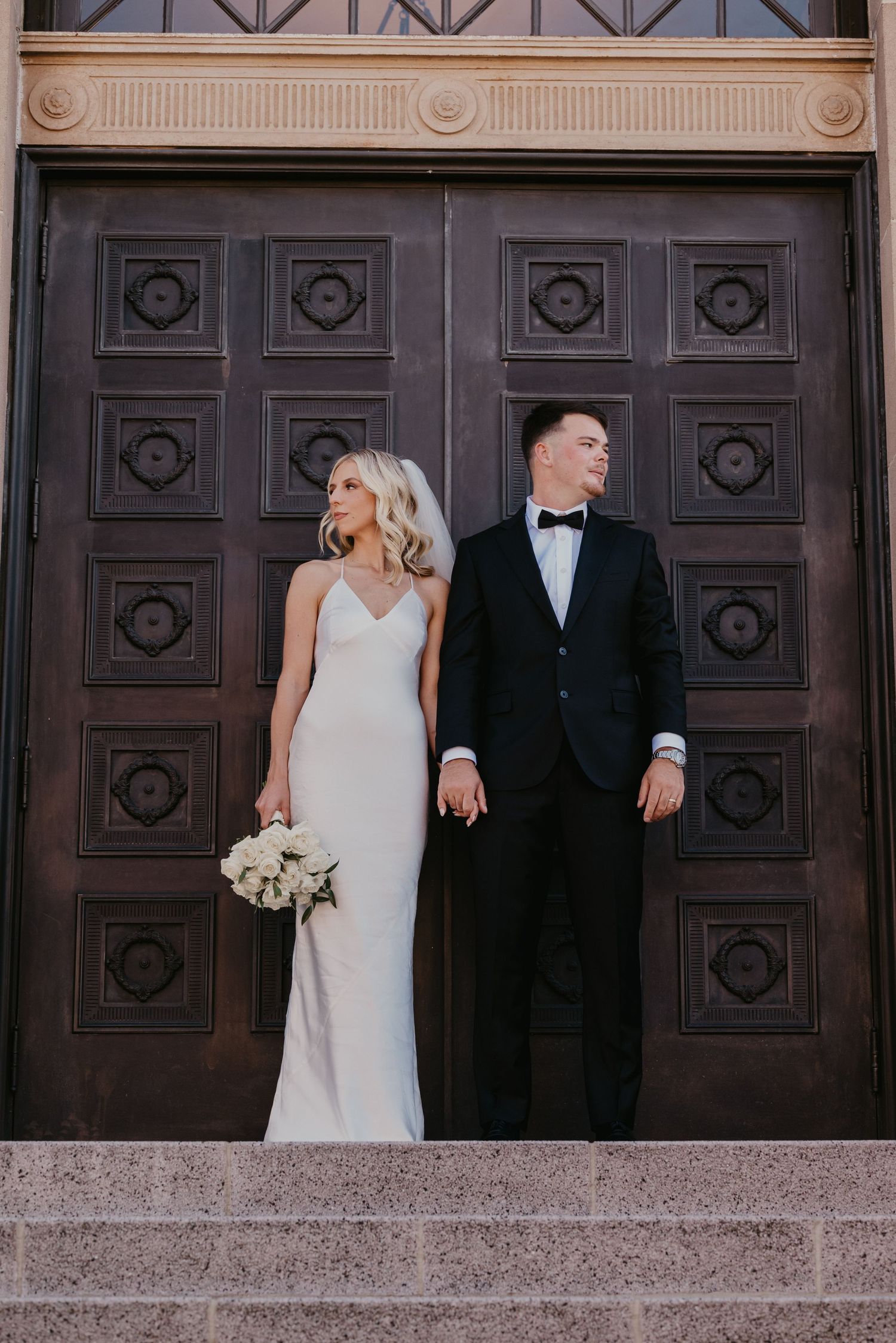 Elegant couple poses in front of ornate wooden doors on their wedding day.