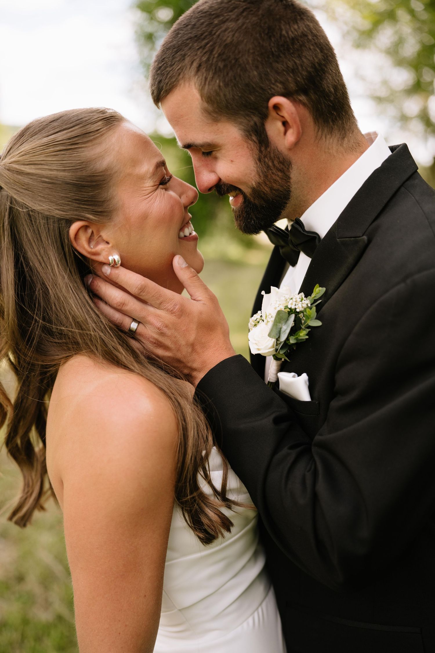 A smiling bride and groom in formal wedding attire share a romantic close moment outdoors, the groom tenderly holding her face.