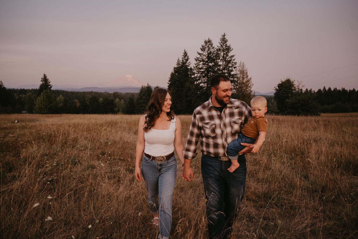 A couple embraces in a golden wheat field during sunset with distant evergreen trees in the background.