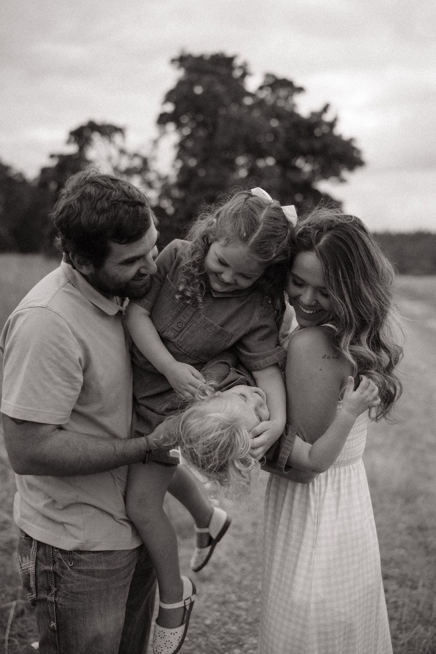 Black and white photo of a happy family with two young daughters laughing together in an outdoor field setting.