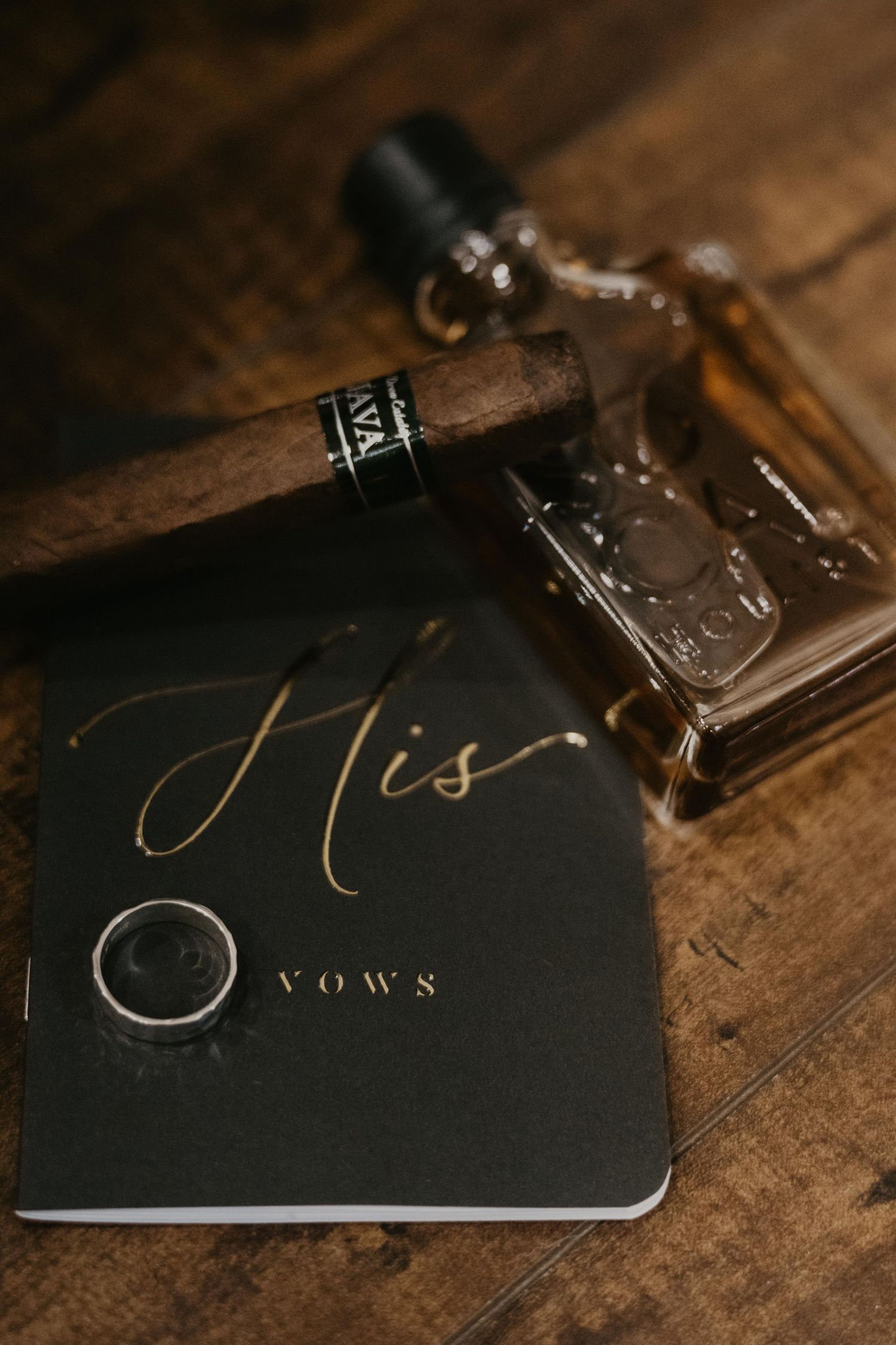 Black His Vows book with gold lettering, wedding ring, cigar, and cologne bottle on rustic wood surface.
