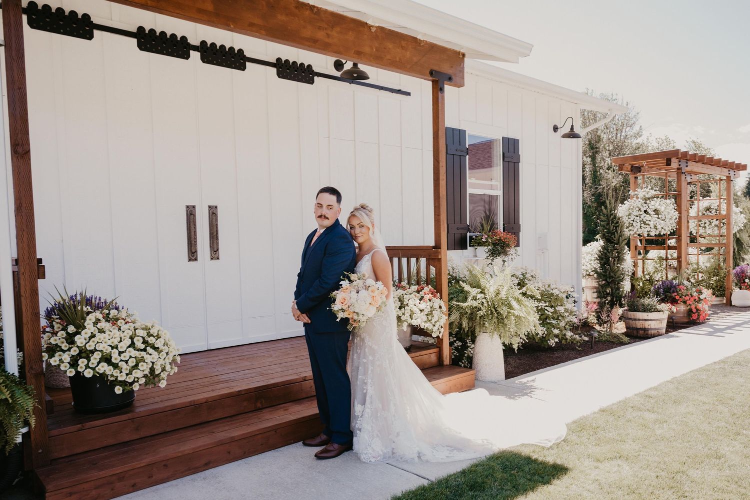 A couple stands together on a wooden deck outside a white barn venue decorated with elegant floral arrangements.