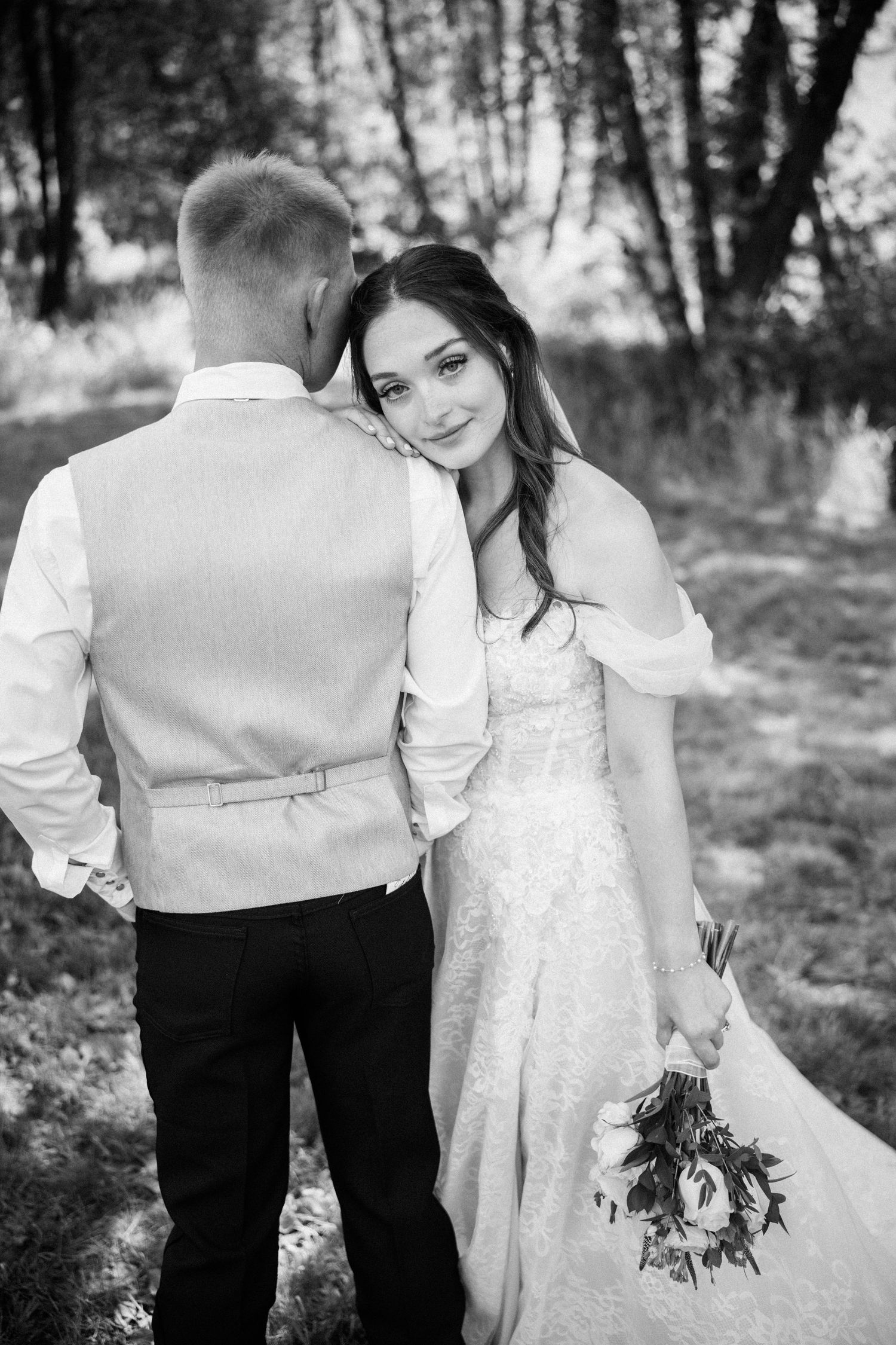 Black and white wedding photo of bride leaning on grooms shoulder holding bouquet outdoors.