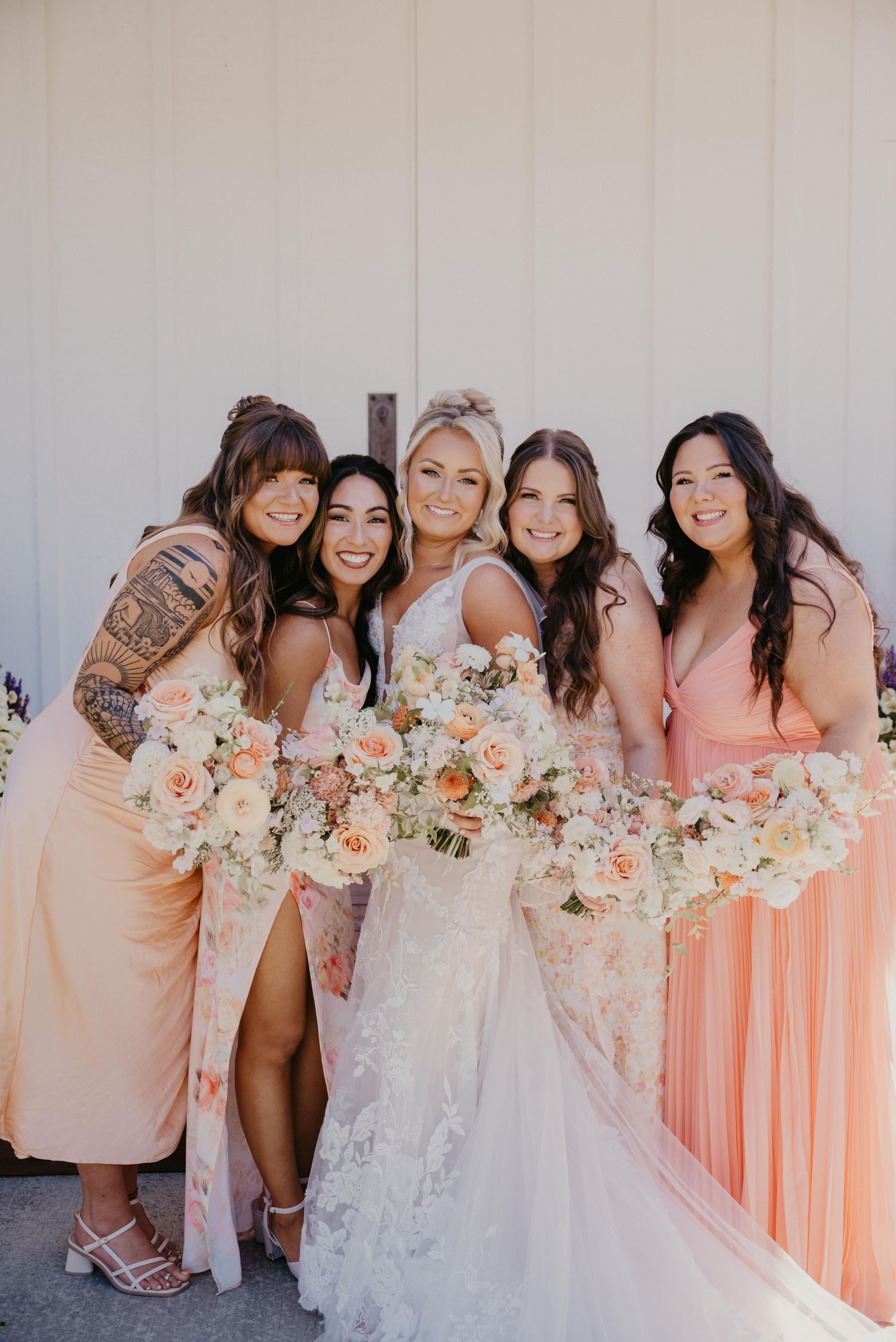 A bridal party in blush pink dresses poses with flower bouquets featuring white and peach blooms against a light wall.