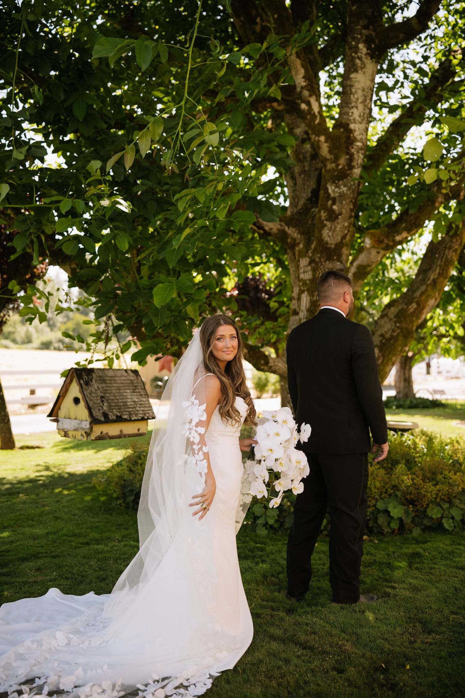 Bride in a lace wedding gown with a long veil holds a white floral bouquet beside a groom in a black suit under a leafy tree.