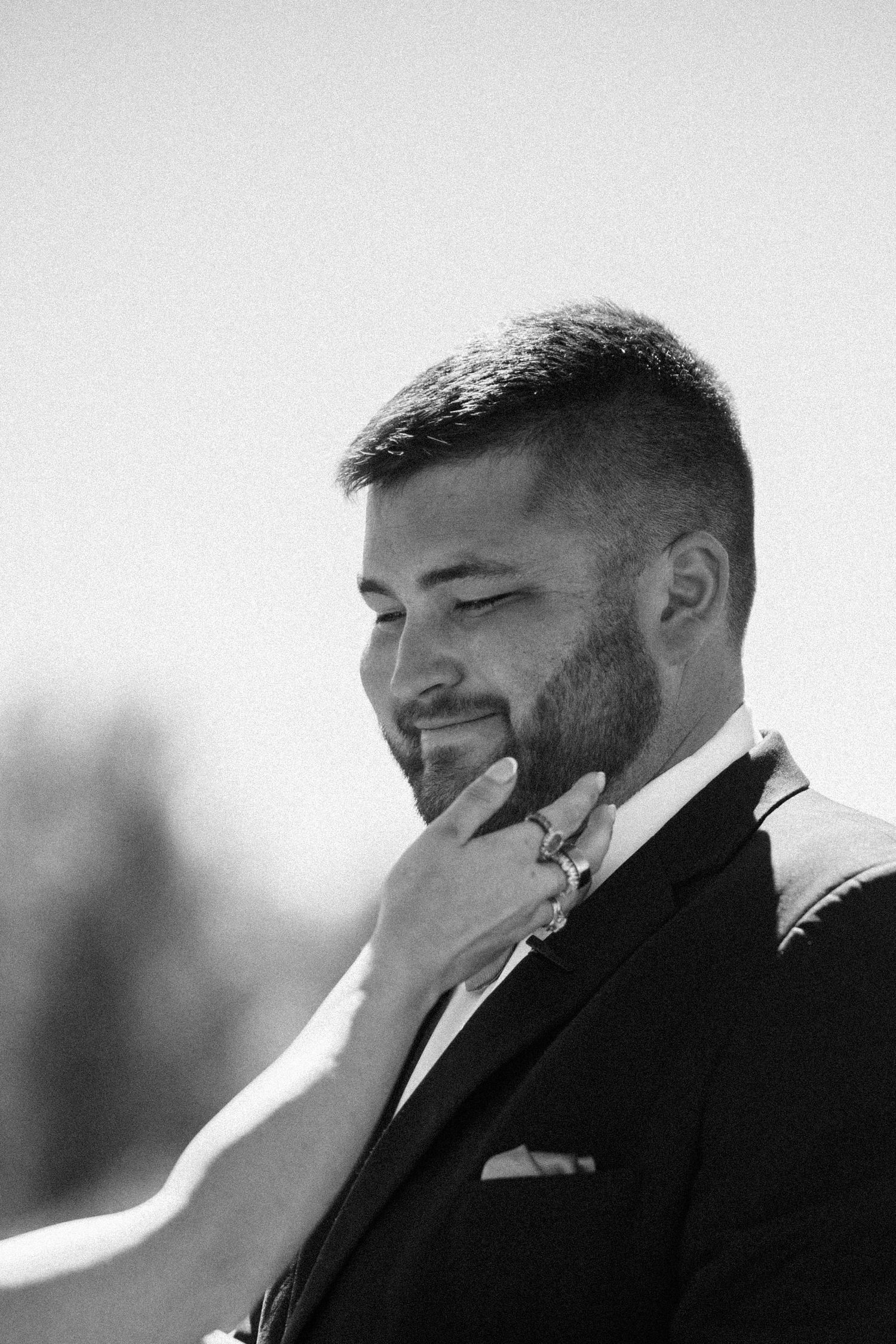 Black and white portrait of a smiling bearded groom in a suit and pocket square as a ring-wearing hand gently touches his chin.