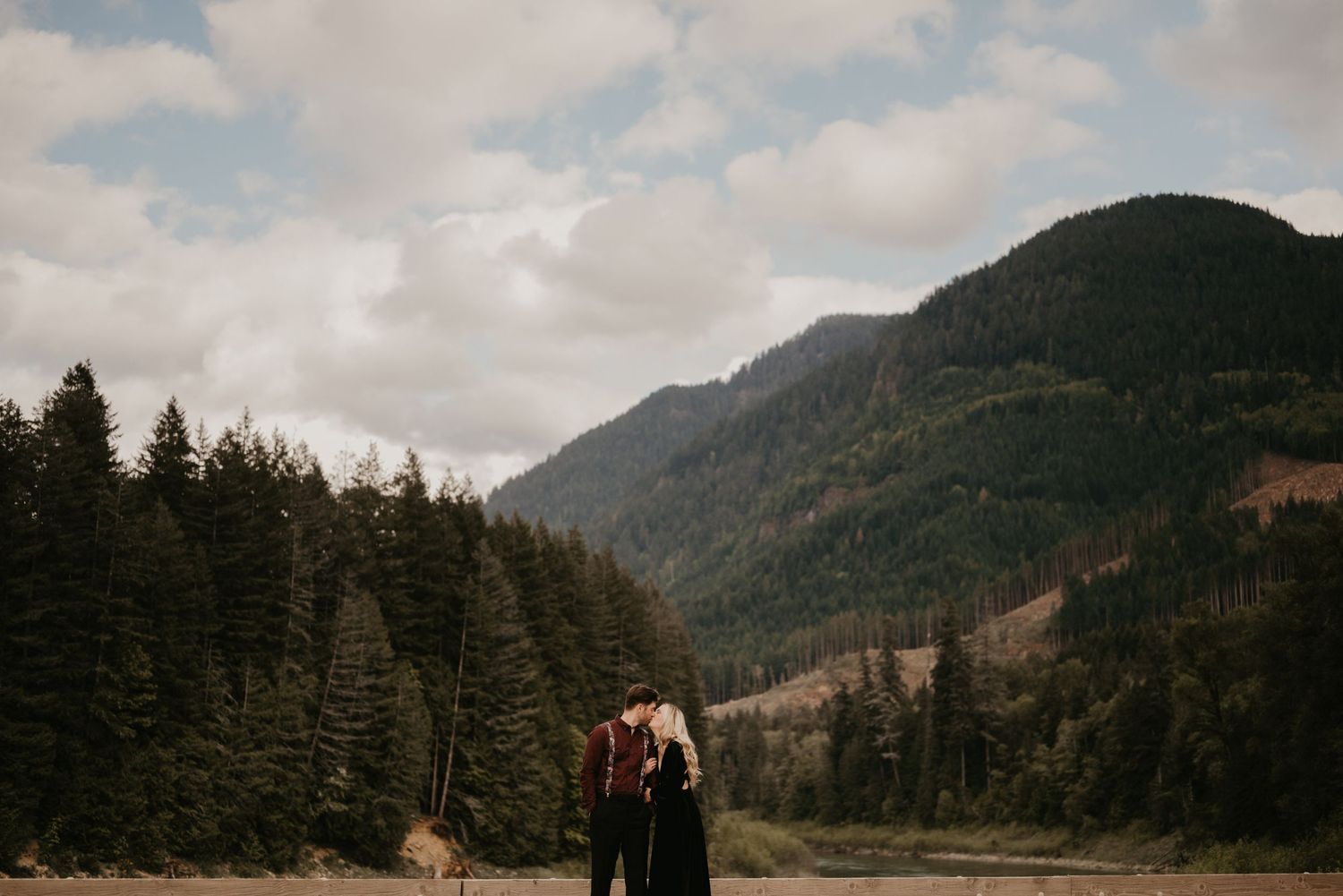 A couple stands in a mountain valley surrounded by evergreen forest and cloudy skies.