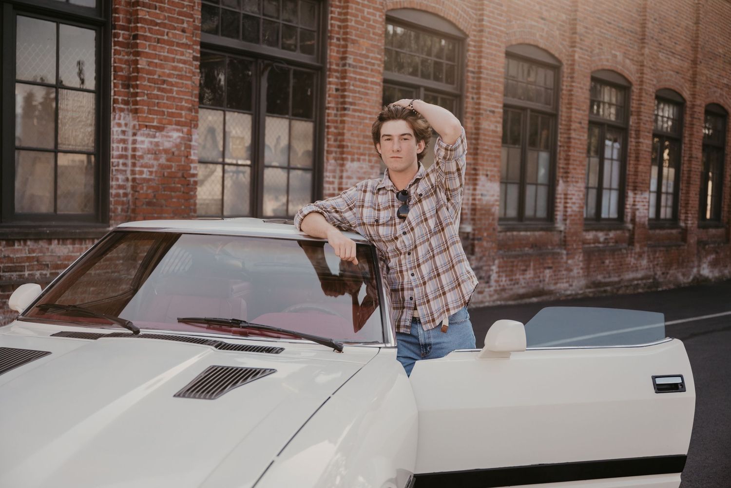Person in plaid shirt leans against white vintage car outside brick building.