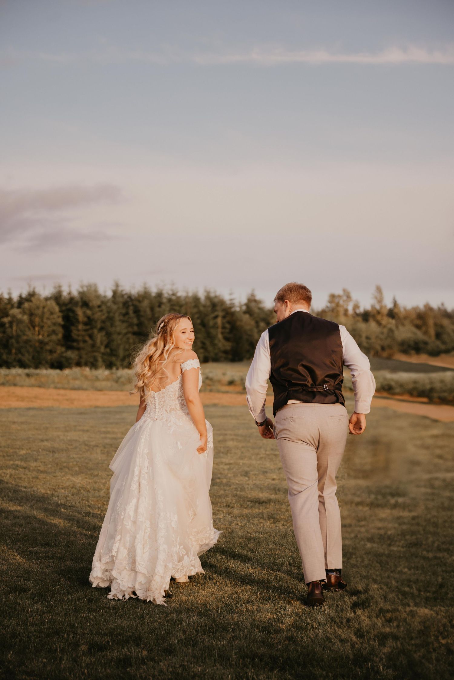 A bride in a lace wedding dress and groom in vest walk together on grassy field at golden hour.