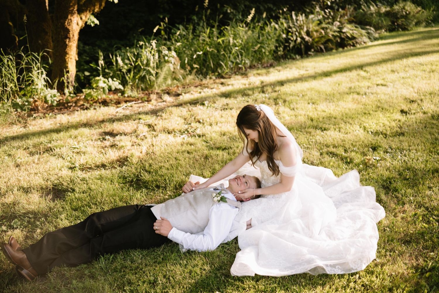 Bride in white gown sits on grass with groom lying down, sharing a romantic moment outdoors on their wedding day.