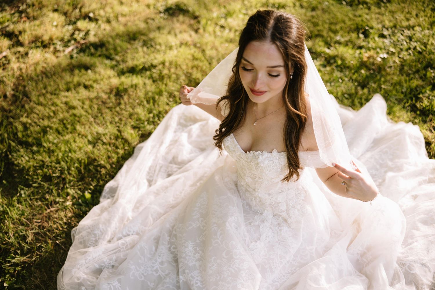A bride in a flowing white lace wedding gown sits gracefully on green grass, her veil cascading softly around her.