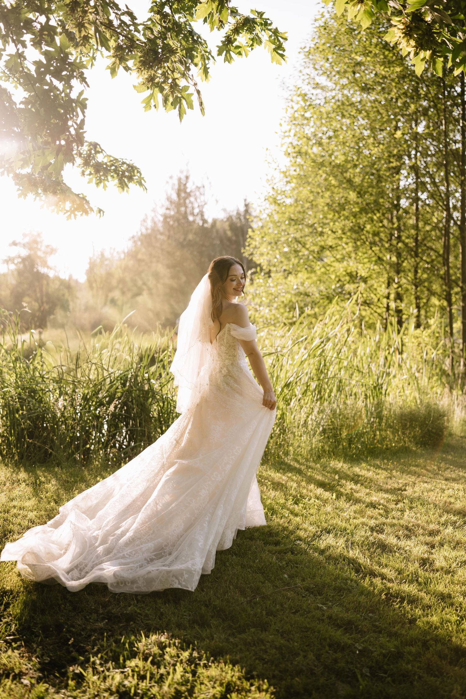Bride in white lace gown and veil stands in sunlit garden surrounded by green trees and tall grass.