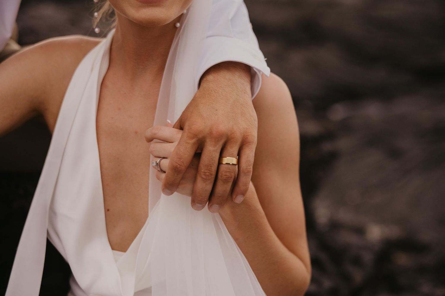 Close-up of a person's hand with a gold wedding band resting on the shoulder of a woman in a white plunging dress against dark rocks.