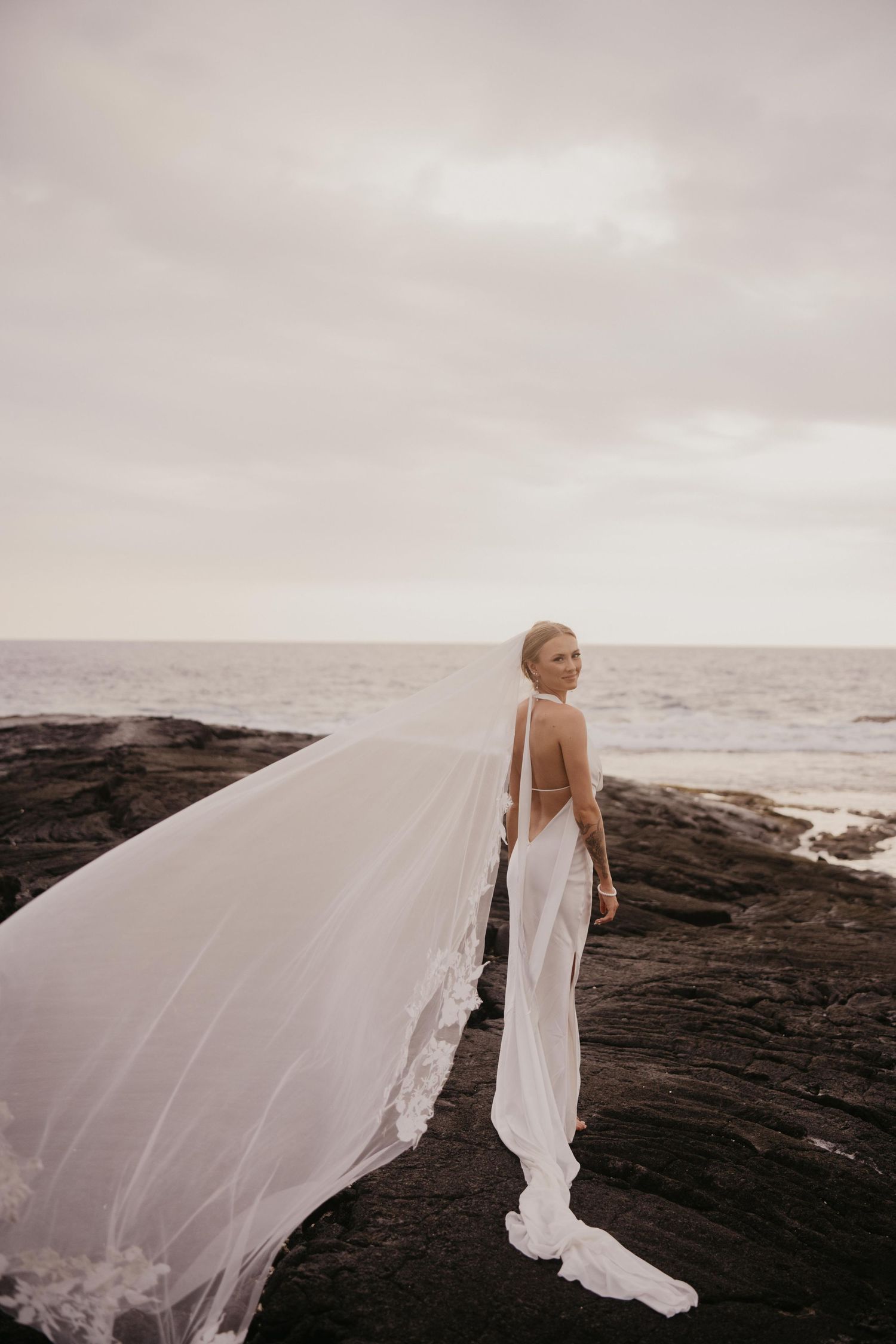 Bride in a backless white gown with a long lace-trimmed veil blowing in the wind, standing on dark volcanic rocks by the ocean.