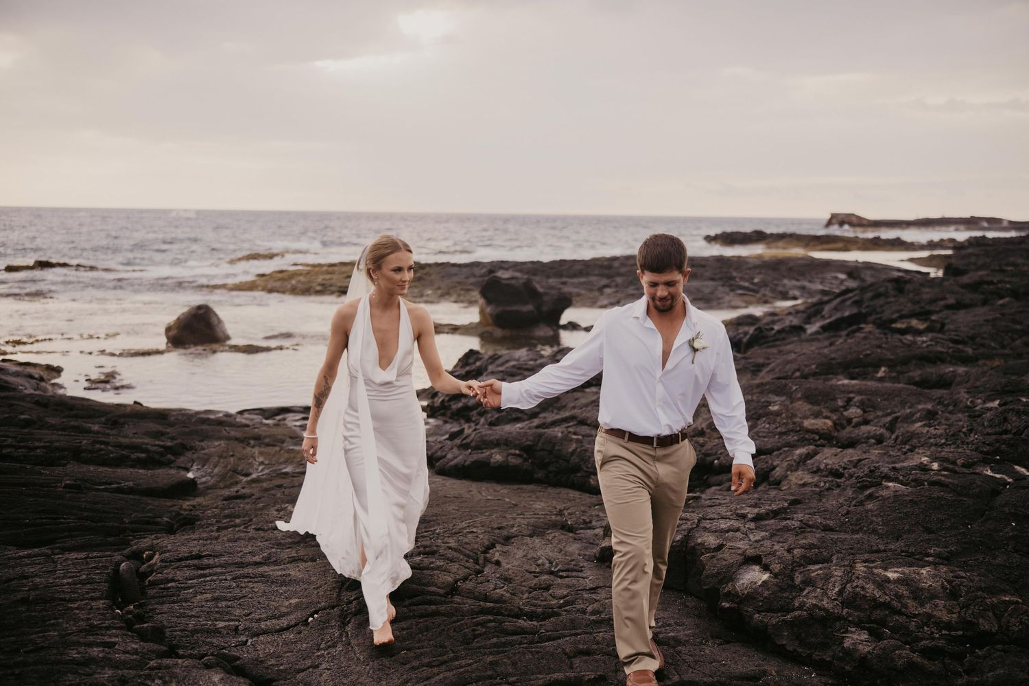 Bride in a flowing white dress holds hands with a groom in a white shirt and khaki pants while walking on black lava rocks by the ocean.