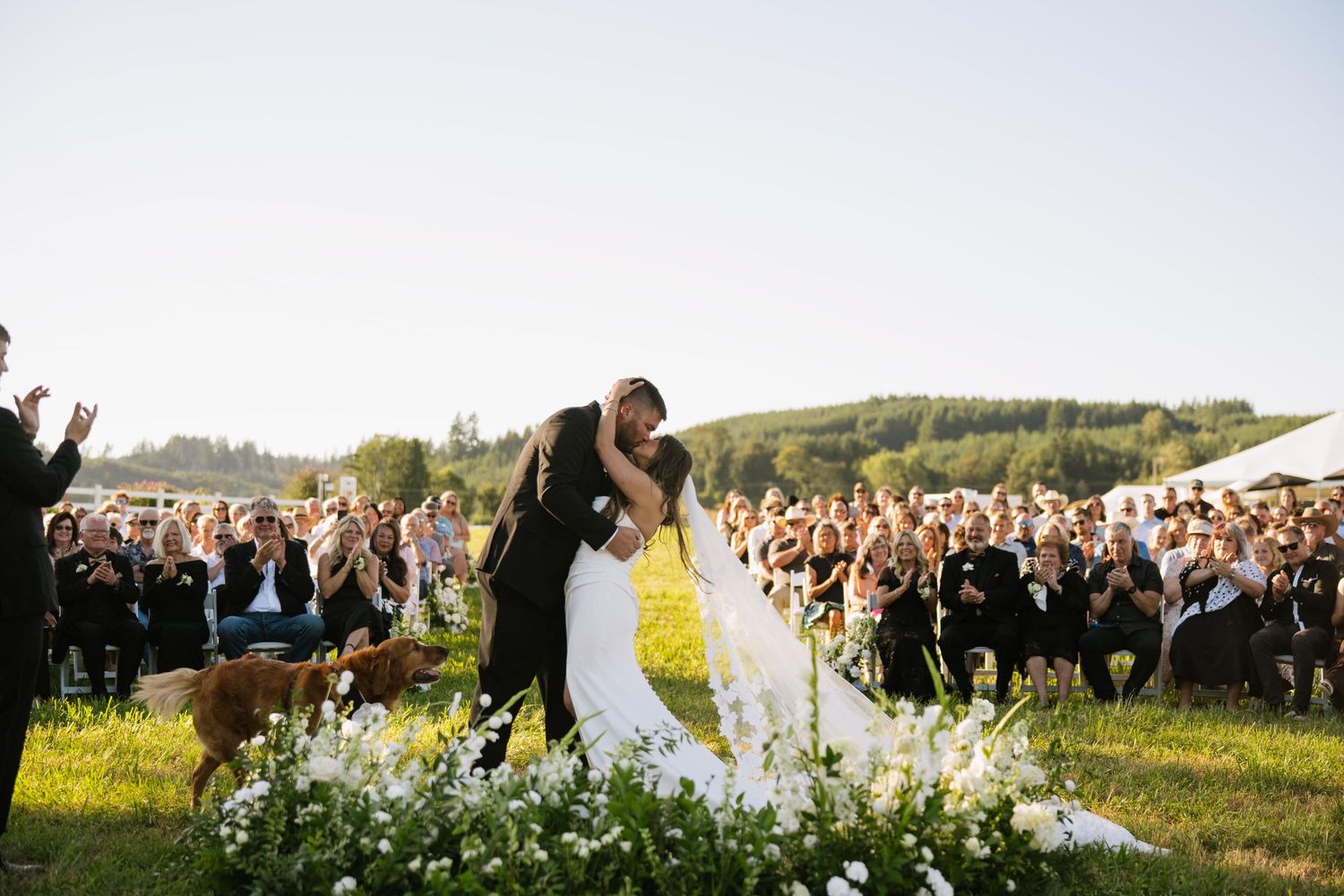 Bride and groom share a kiss during an outdoor wedding ceremony as guests applaud and a dog watches beside a floral arrangement on the lawn.