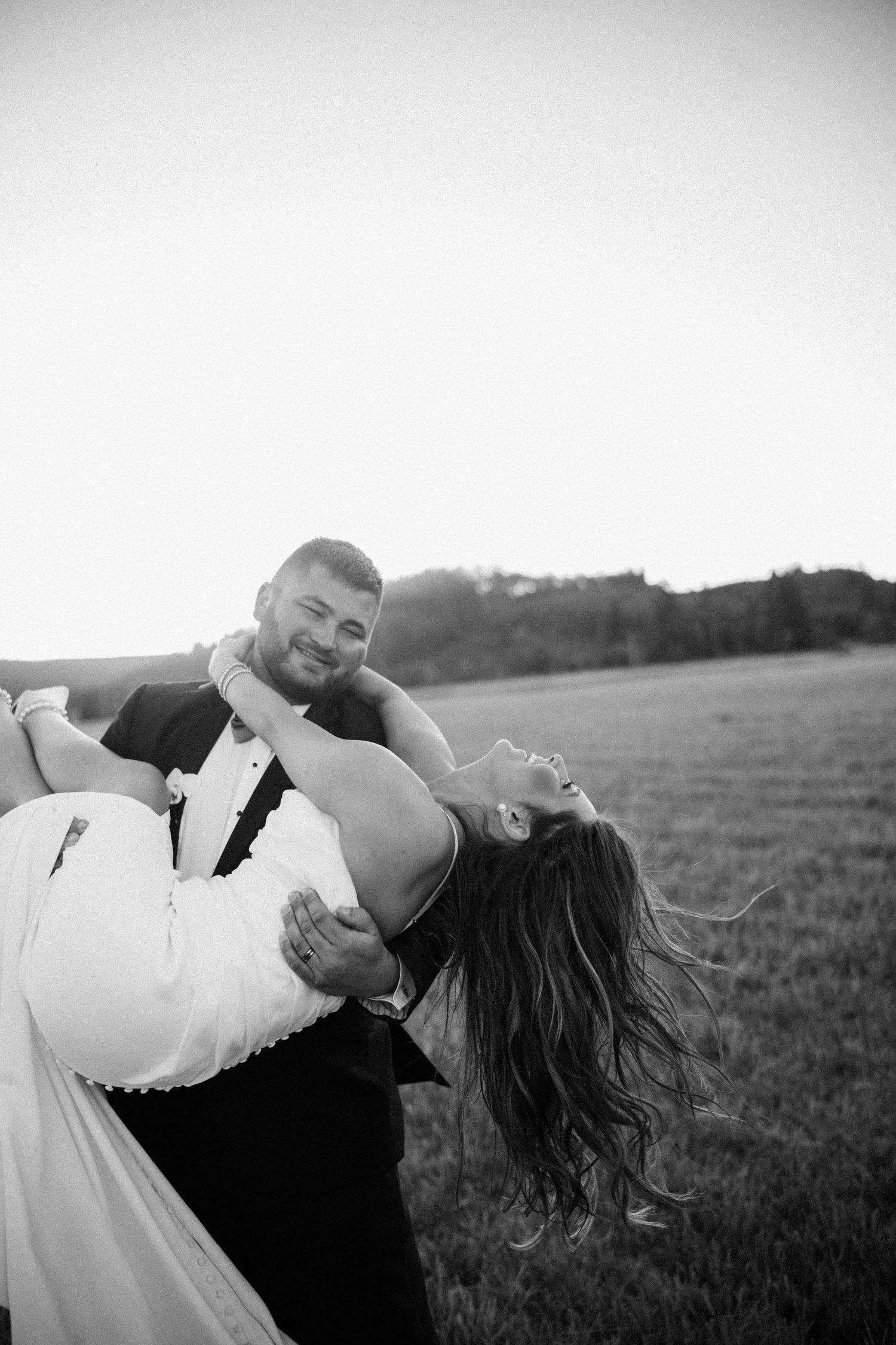 Black and white wedding photo of a smiling groom in a tuxedo cradling his laughing bride in a white gown in a grassy field at sunset.