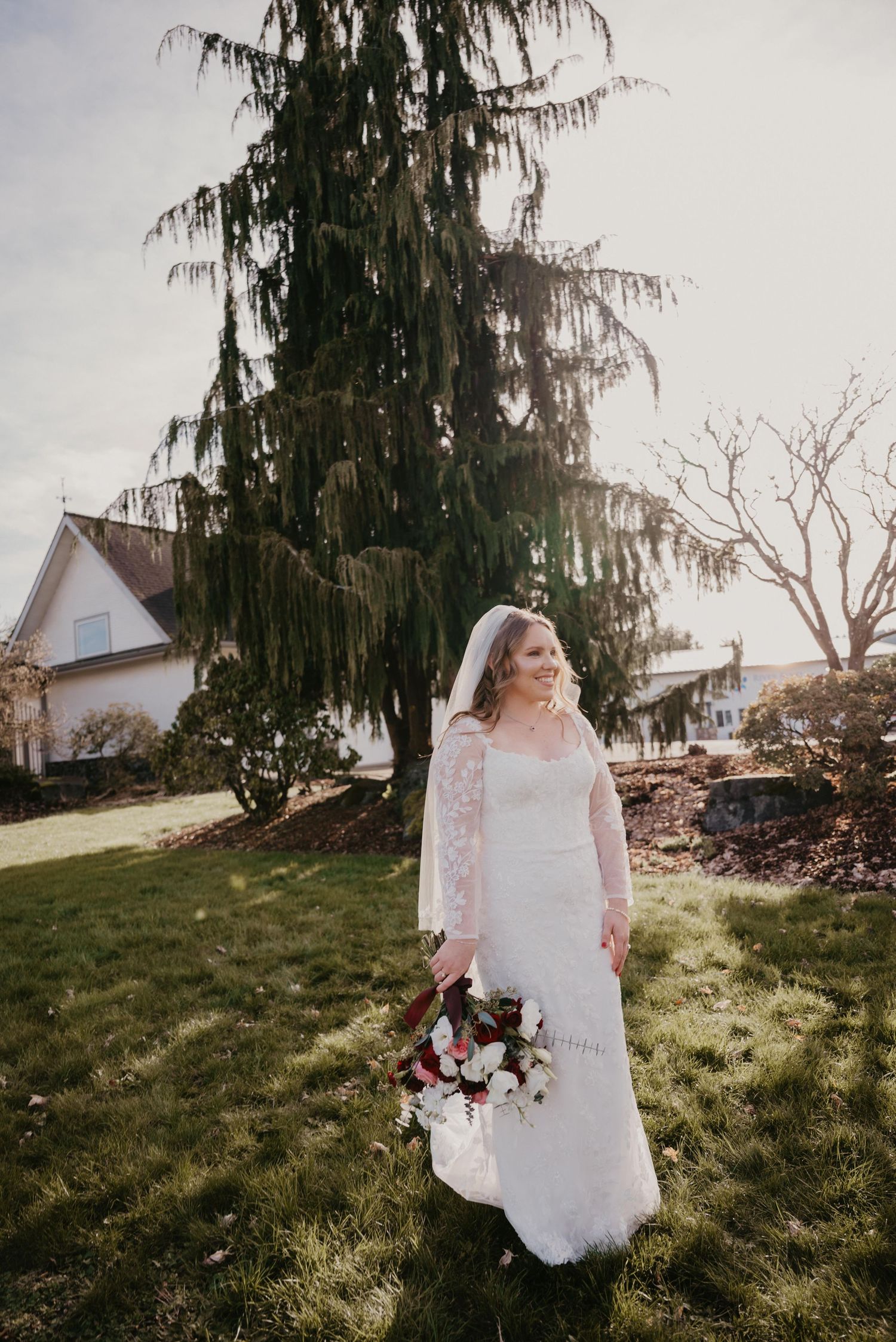 A bride in a white lace gown holds a burgundy bouquet while standing on a grassy hill during golden hour.