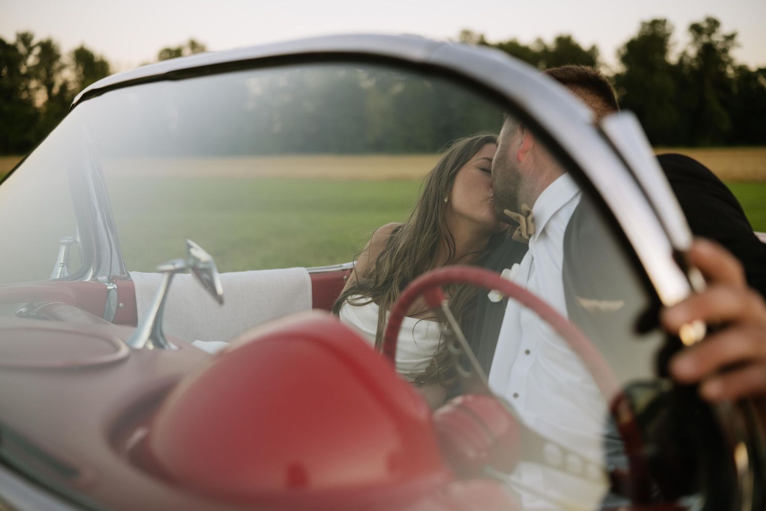 Couple kissing in a vintage red convertible, seen through the windshield, with a sunlit green field and trees in the background.