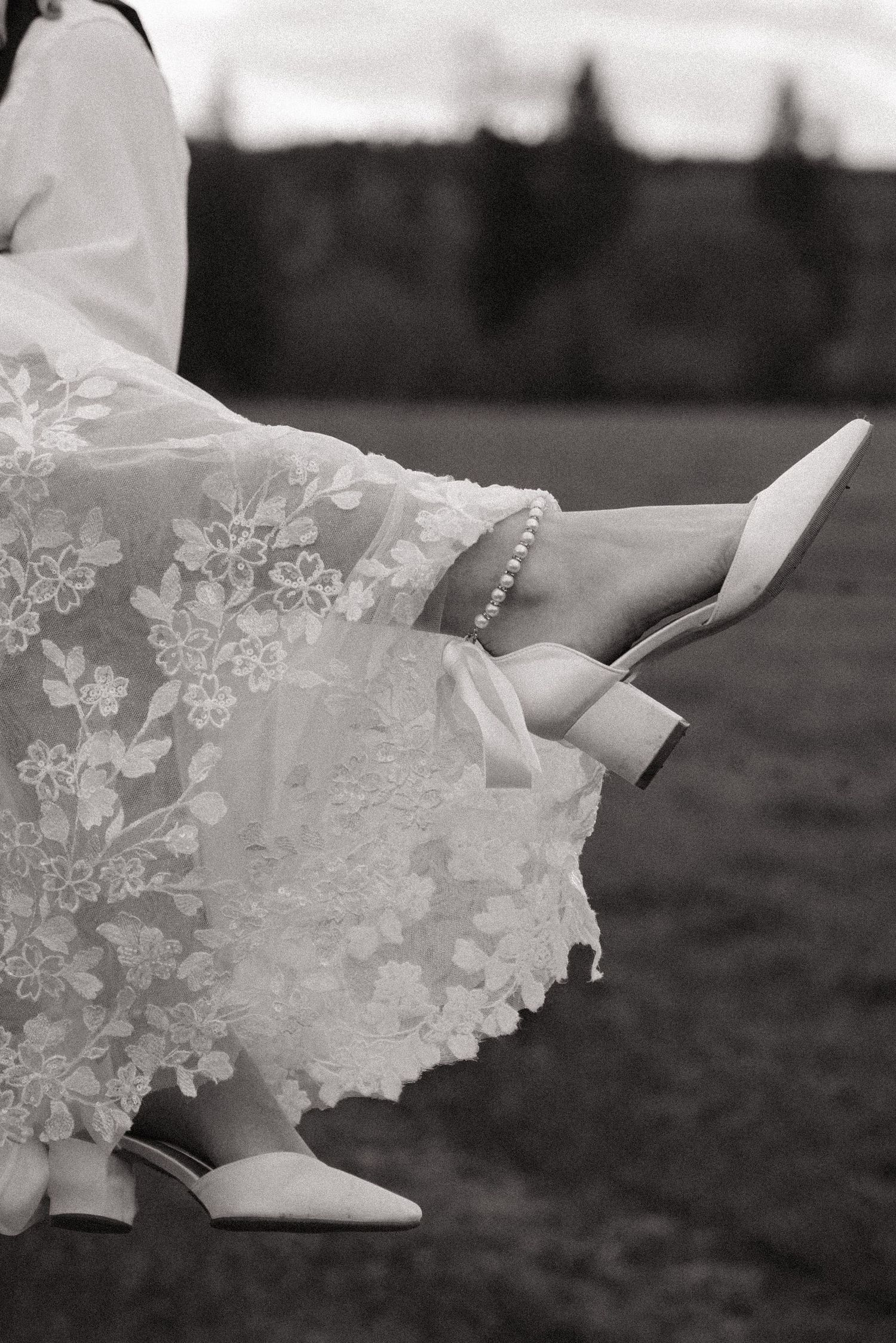 A black and white photograph shows delicate lace details on a wedding dress hem and shoes.