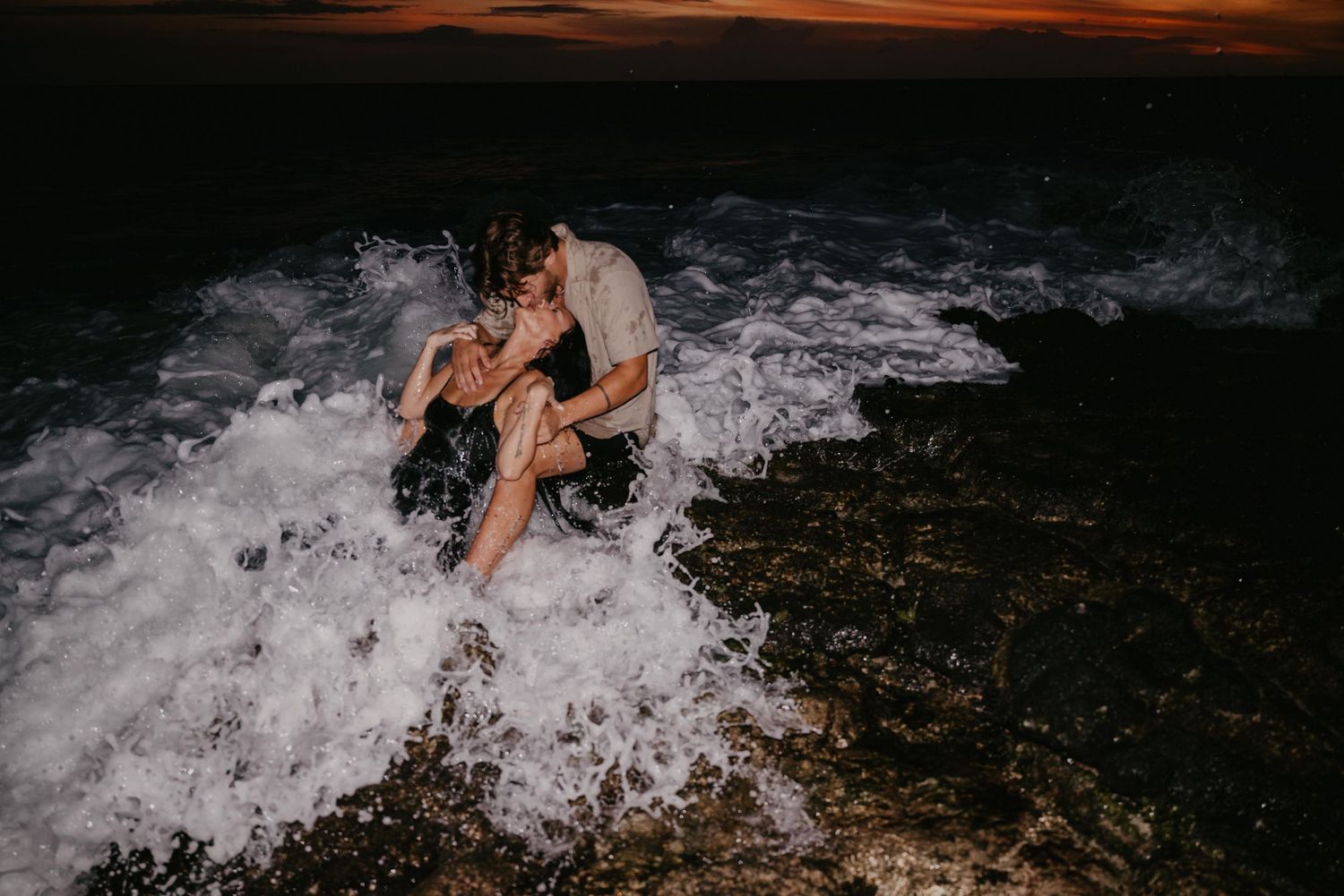 Breaking waves crash against dark volcanic rocks during sunset at the beach, creating dramatic ocean spray.
