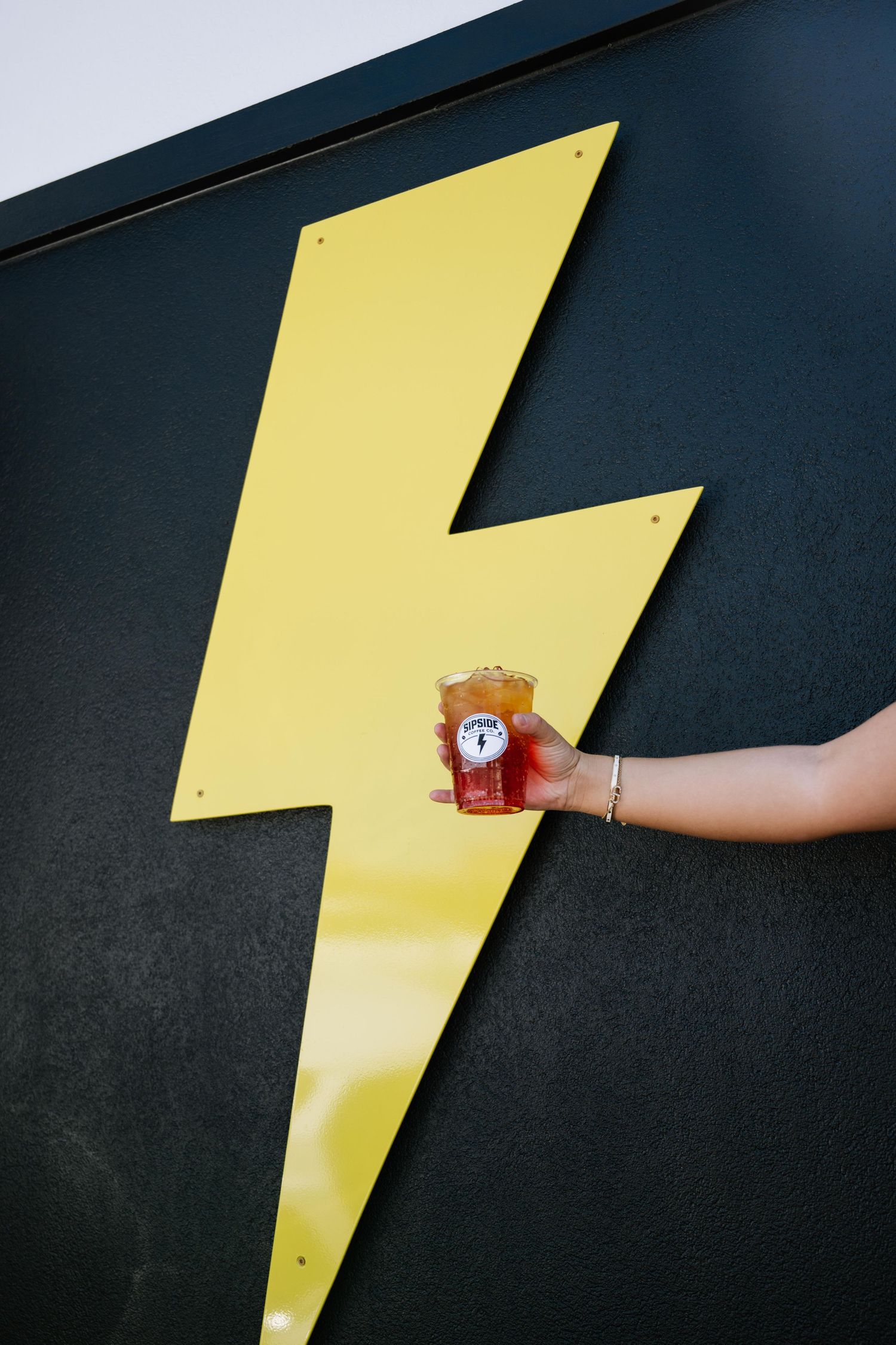 A hand holds an iced drink in front of a bold yellow lightning bolt sign on a dark wall.