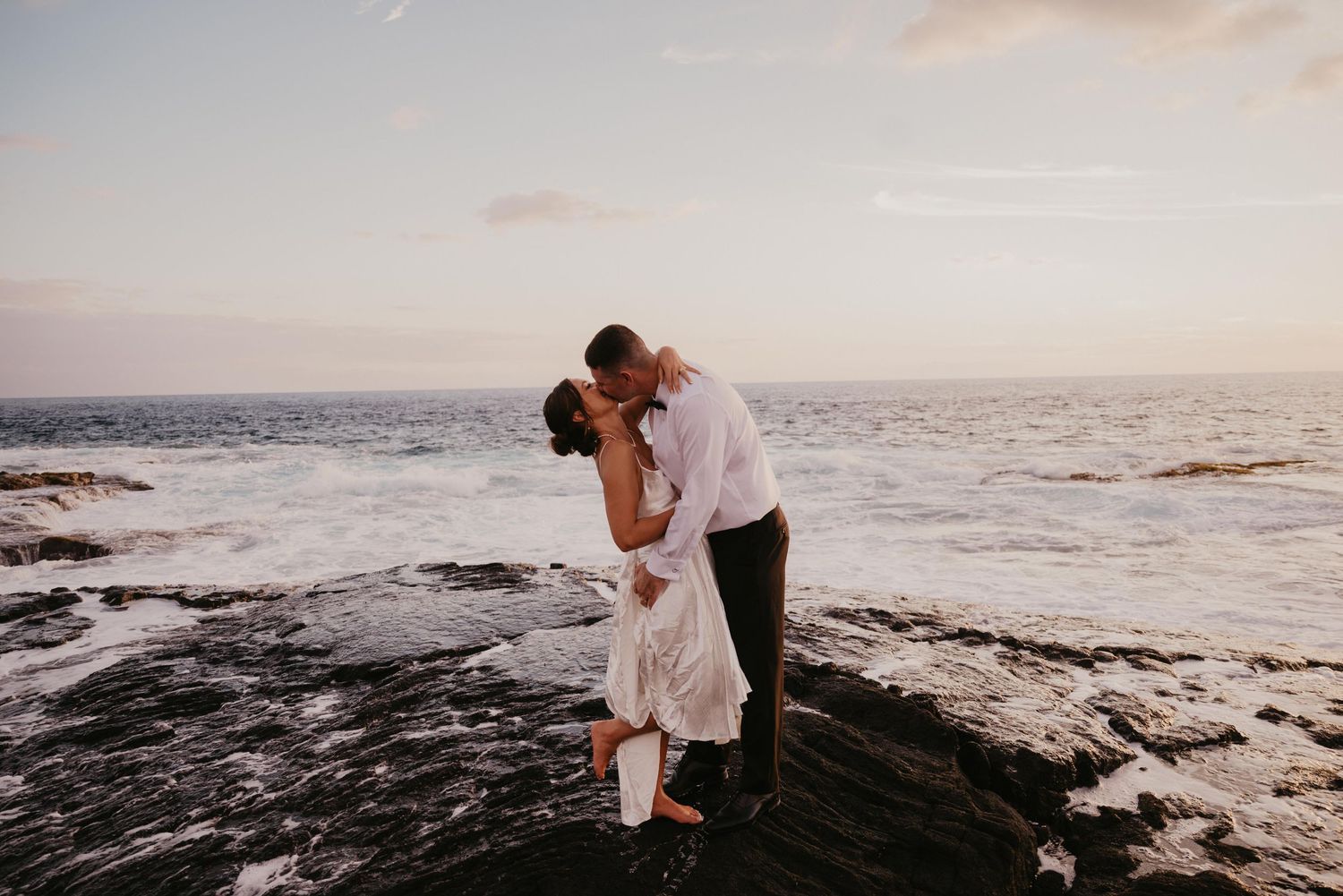 Couple embracing on rocky beach at sunset with ocean waves crashing in the background.