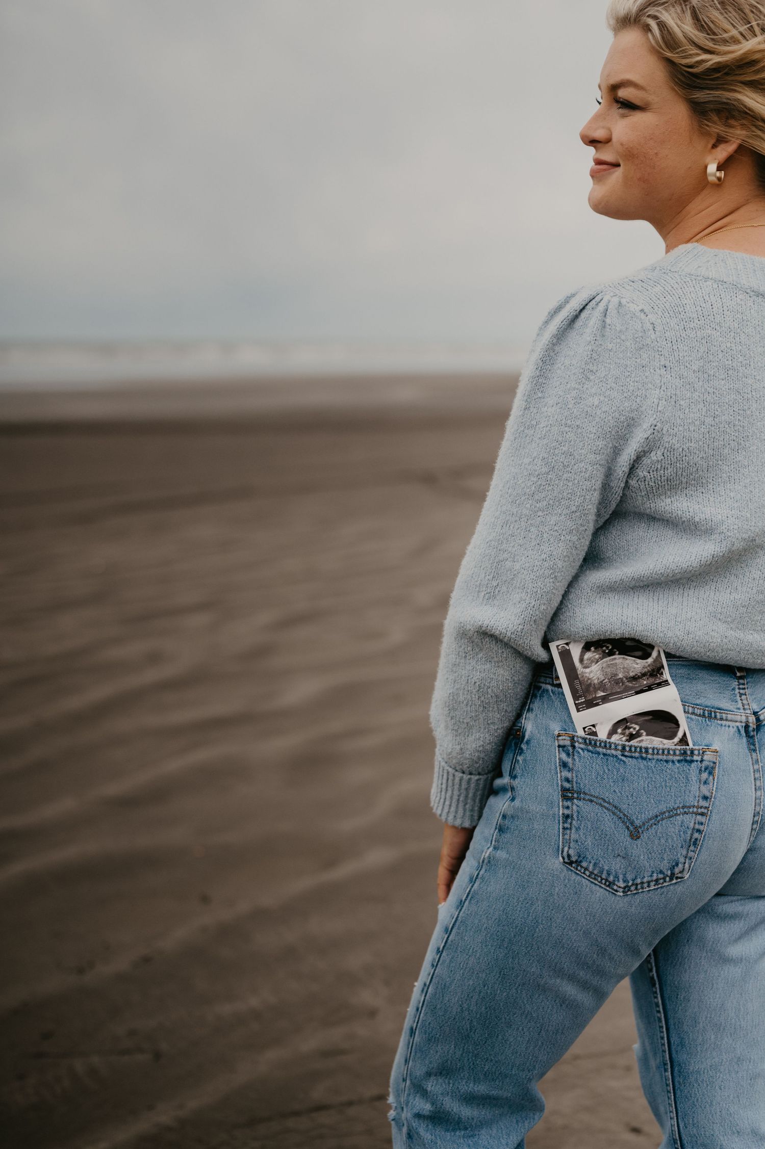 Person in grey sweater and jeans standing on beach with ultrasound photo in back pocket.