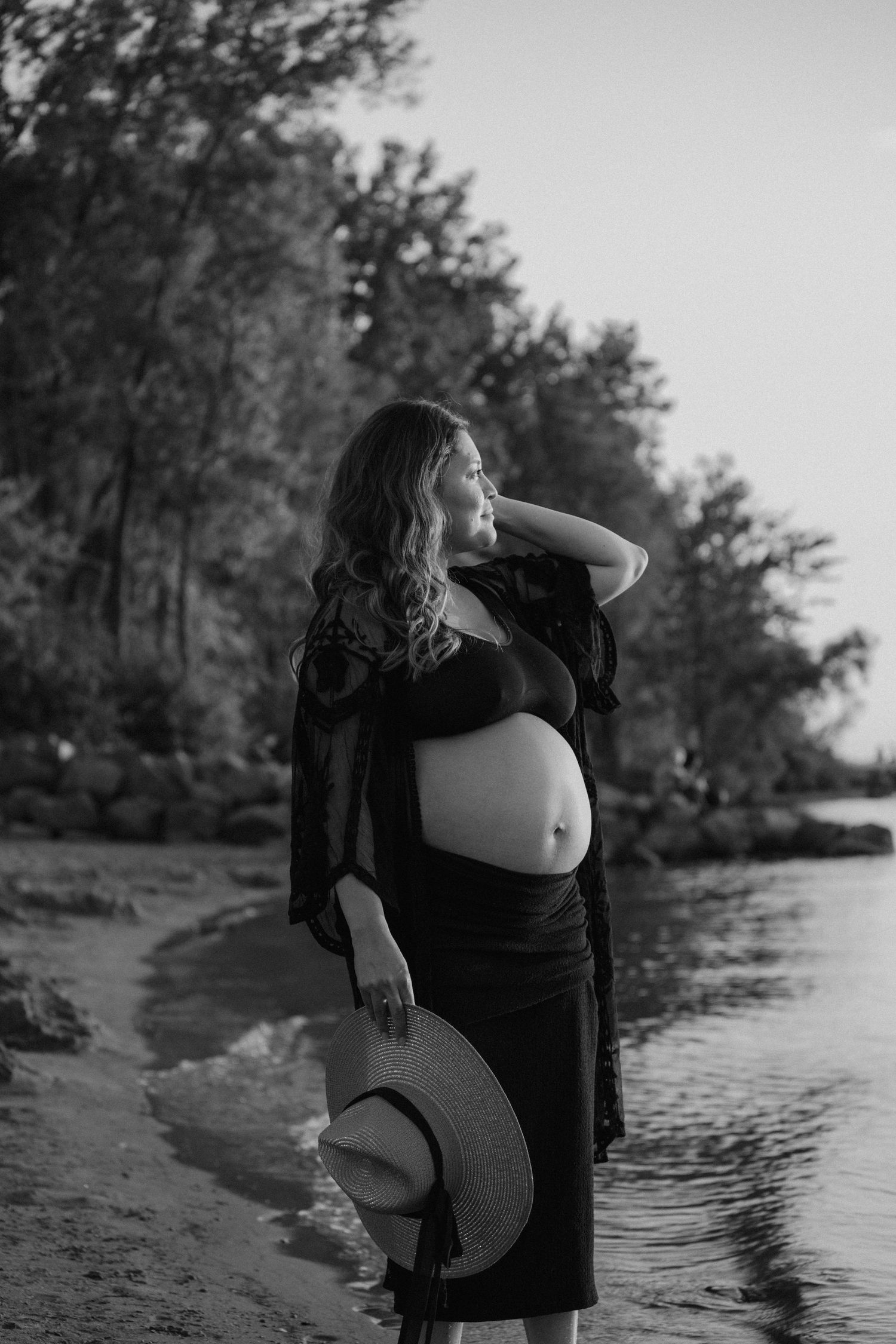 Silhouette of an expectant mother holding a hat while standing on a beach at sunset.