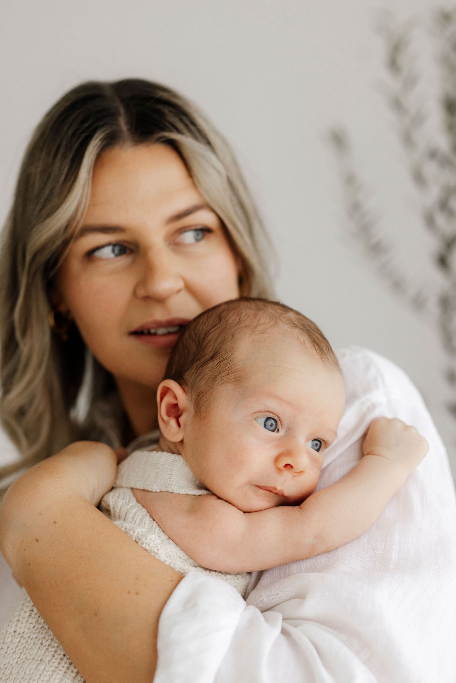 A mother with blonde hair holds her blue-eyed newborn baby against her chest in a bright, airy room.
