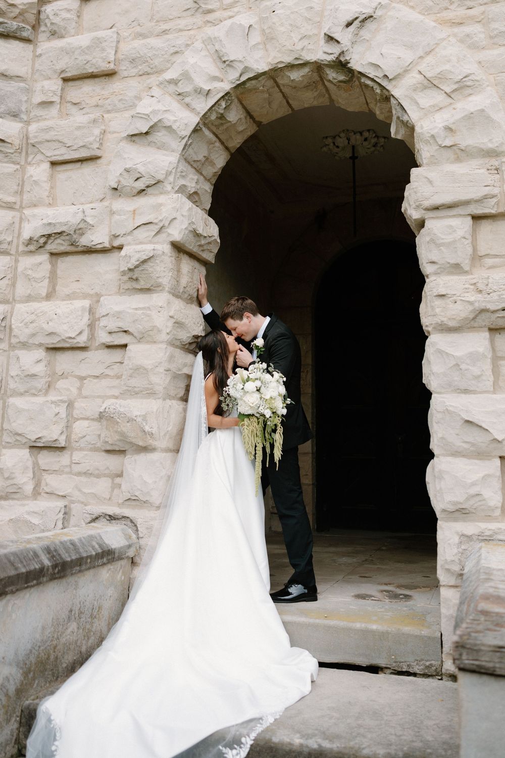 Bride and groom share a romantic kiss under a stone arch, her white gown and bouquet flowing elegantly.