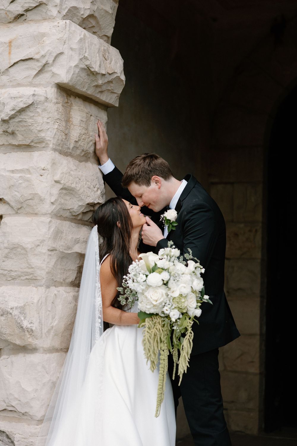 Bride and groom share a romantic kiss against a stone wall, bride holding a lush white floral bouquet with greenery.
