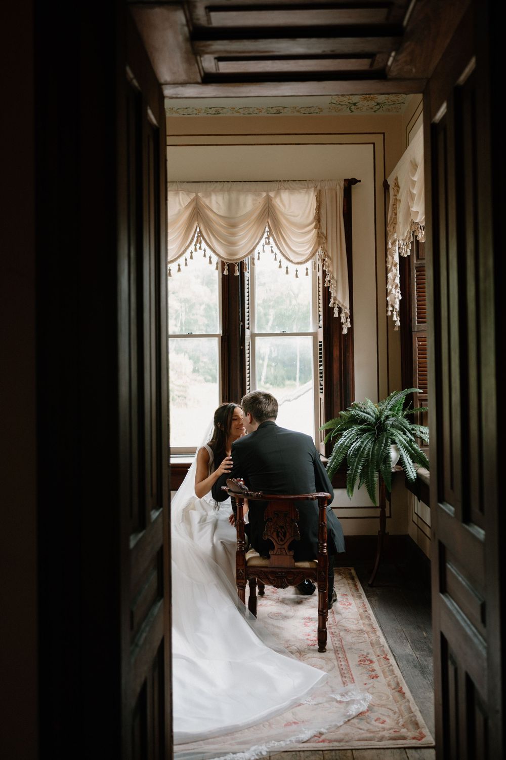 Bride and groom sit intimately by a sunlit window in an elegant vintage room, sharing a quiet moment together.