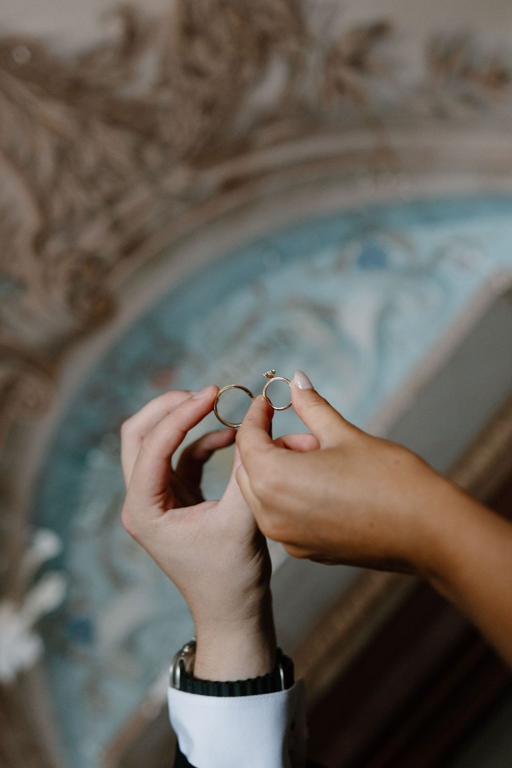 Two hands holding wedding rings together against an ornate decorative background.