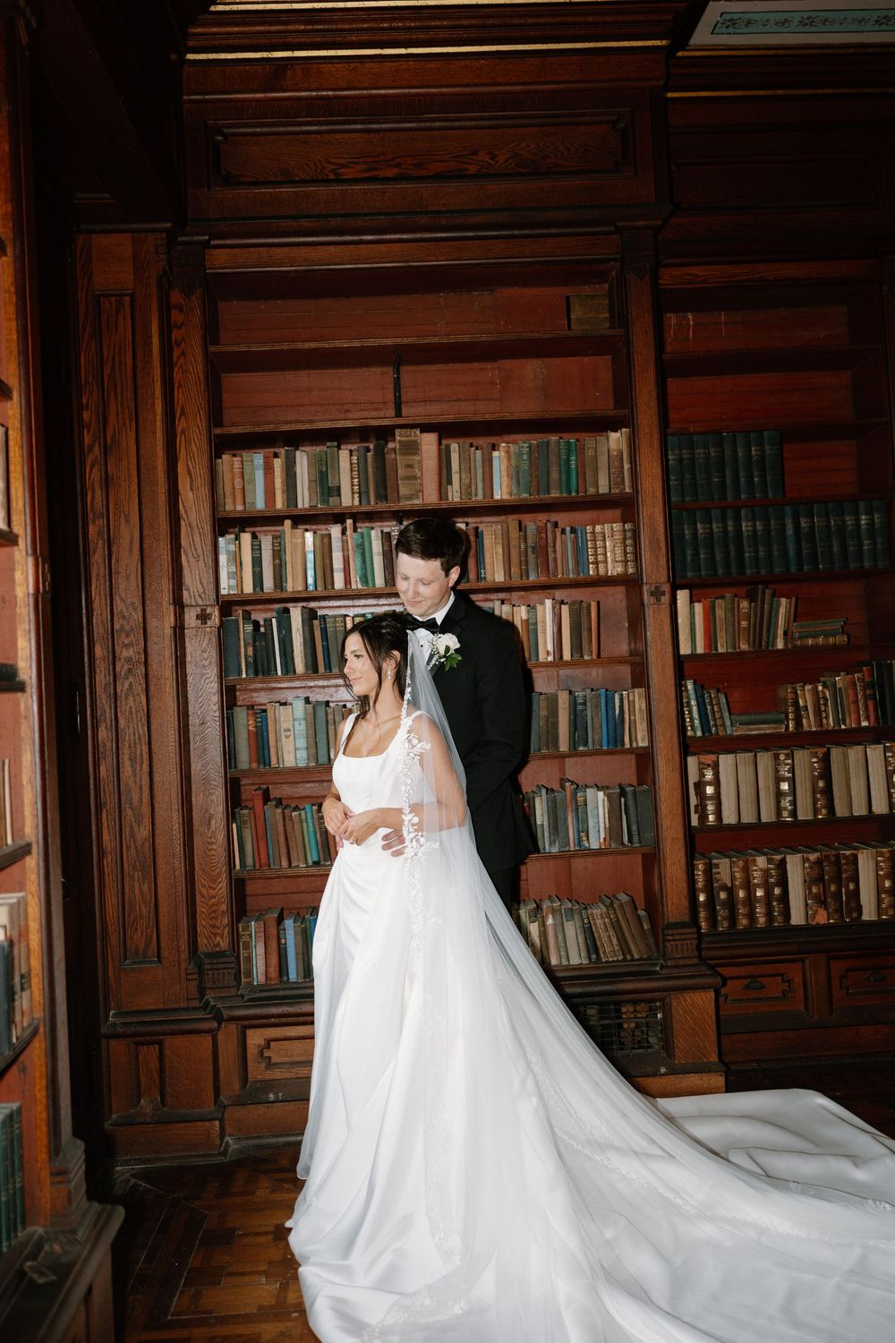 Bride and groom pose romantically in a wood-paneled library filled with vintage books during their wedding.