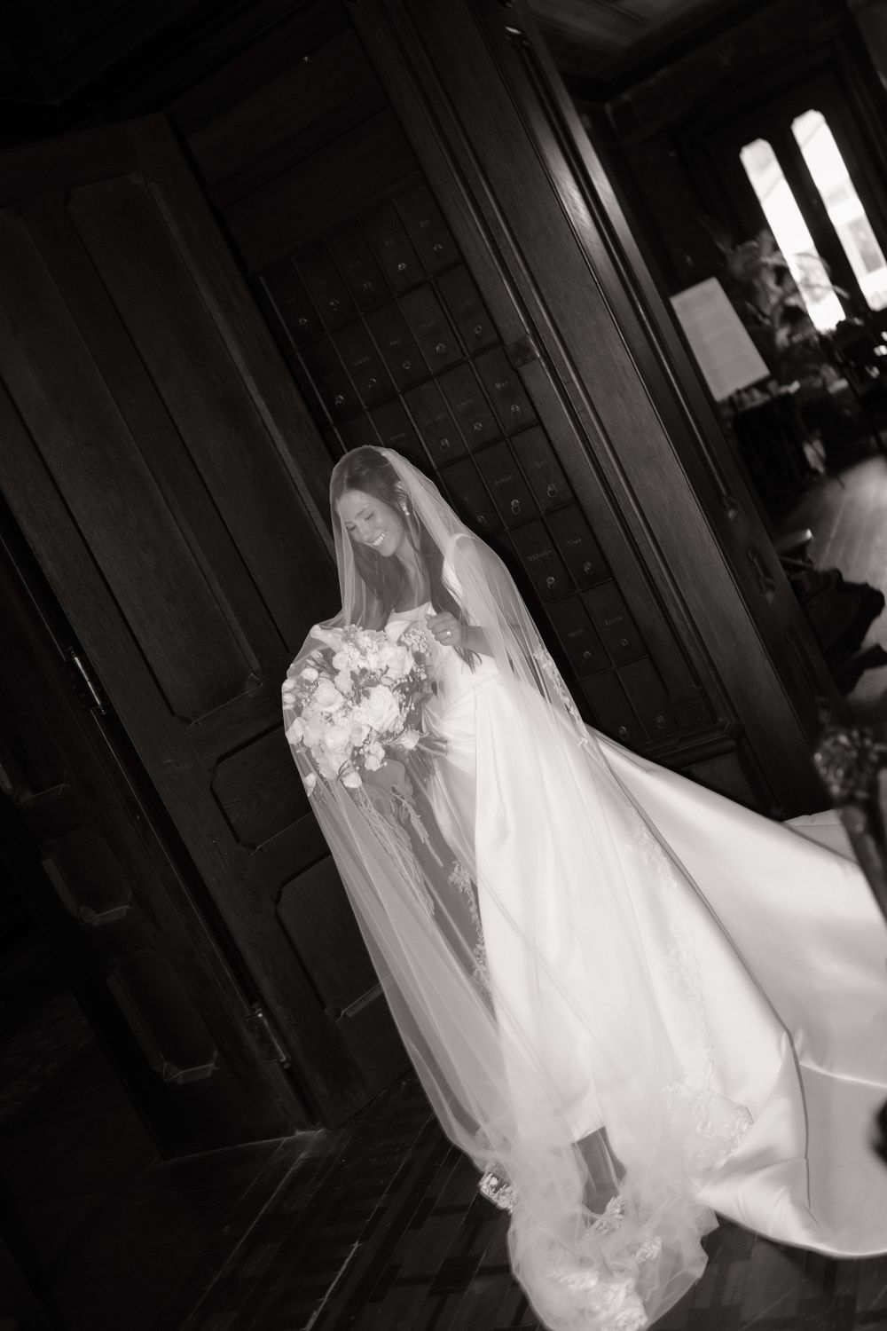 Elegant bride in a flowing white gown and veil holds a bouquet in a black and white wedding portrait.
