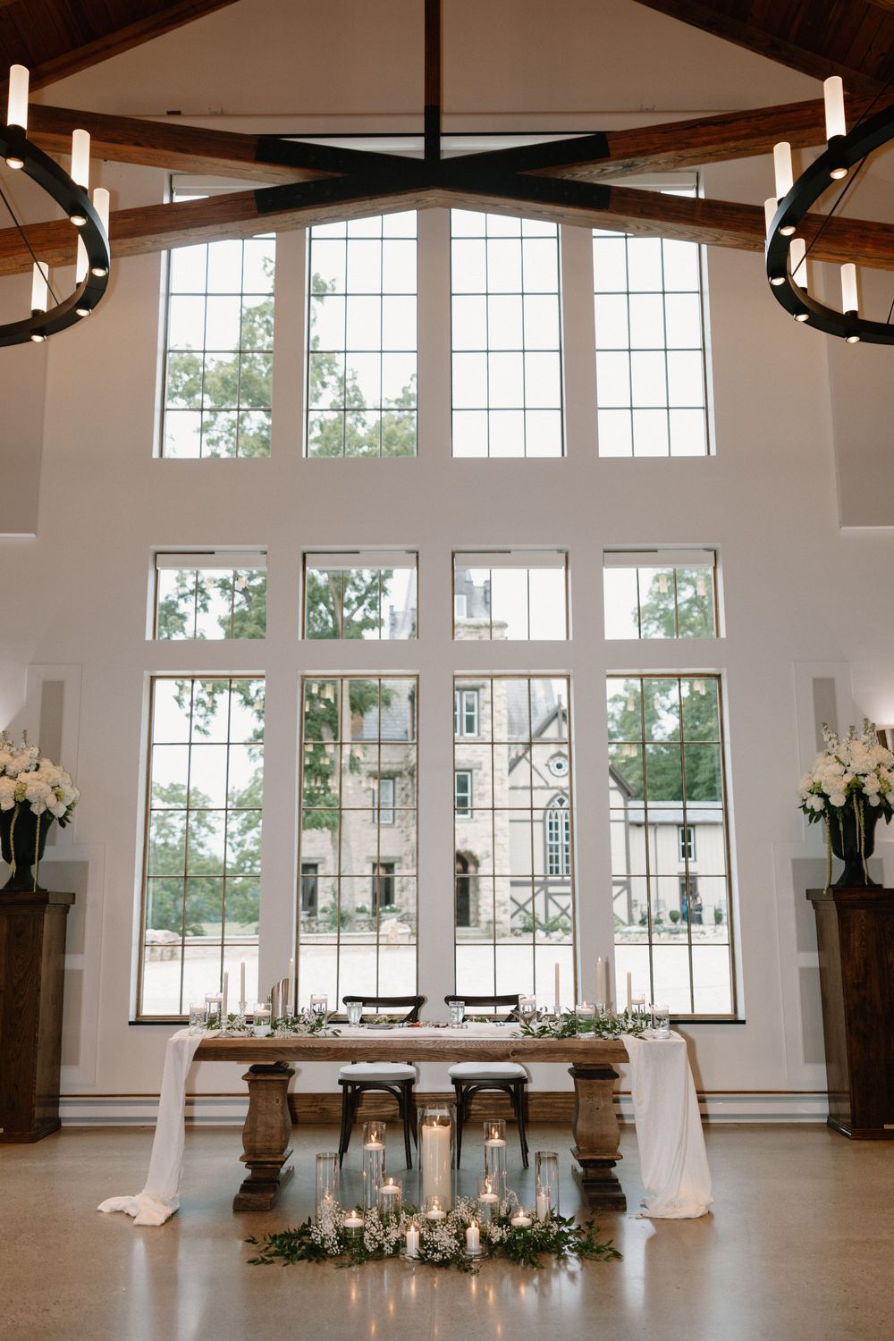 Stunning wedding reception sweetheart table with candles and greenery before tall cathedral windows.