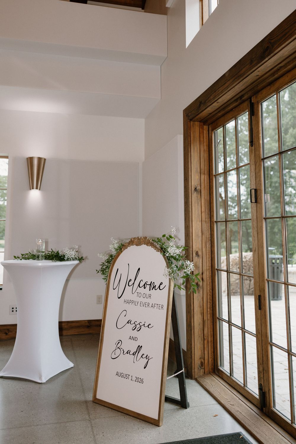 Elegant wedding welcome sign for Cassie and Bradley, August 1, 2026, displayed near glass doors with floral decor.