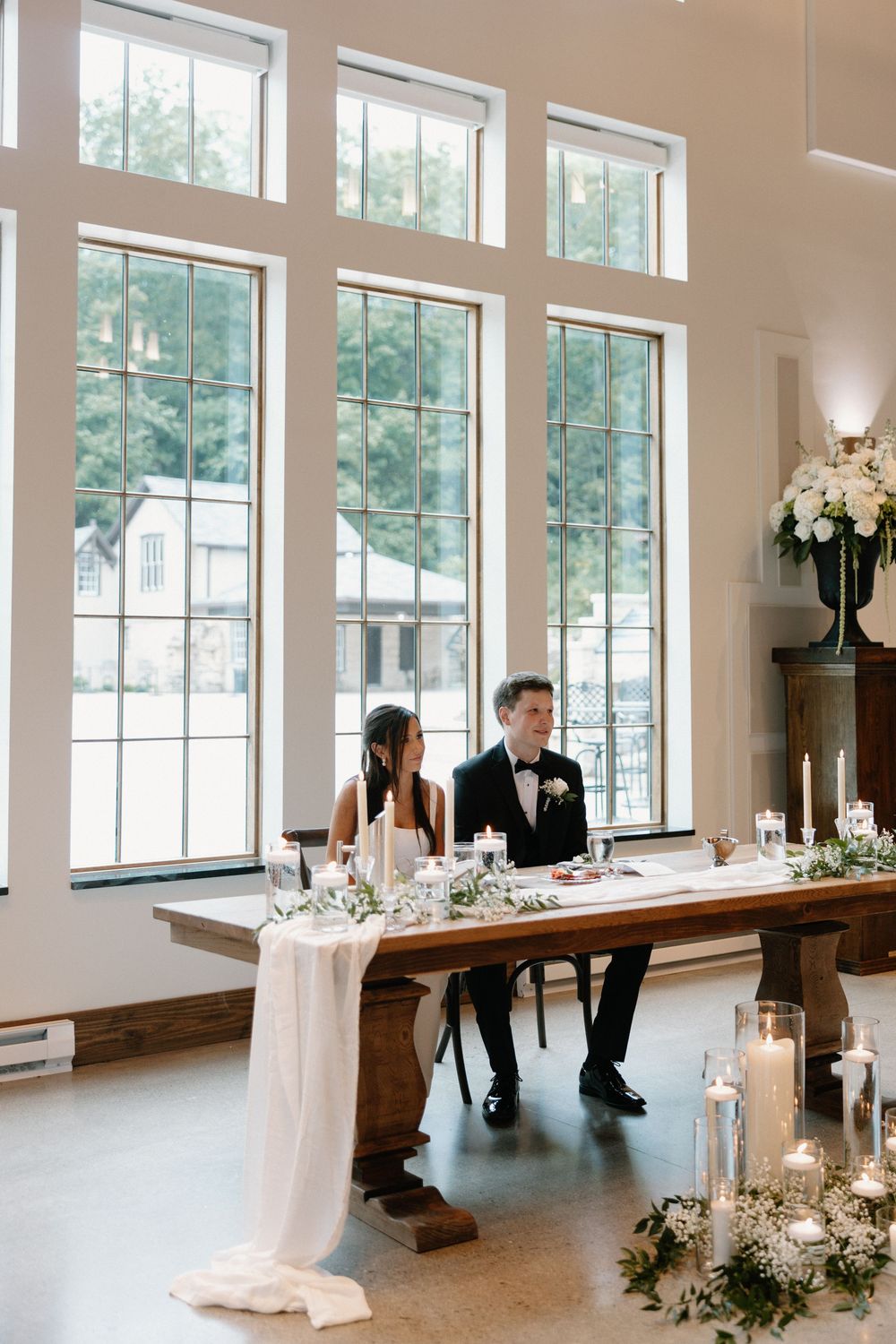 Bride and groom seated at rustic wooden sweetheart table adorned with candles and greenery before tall white-framed windows.