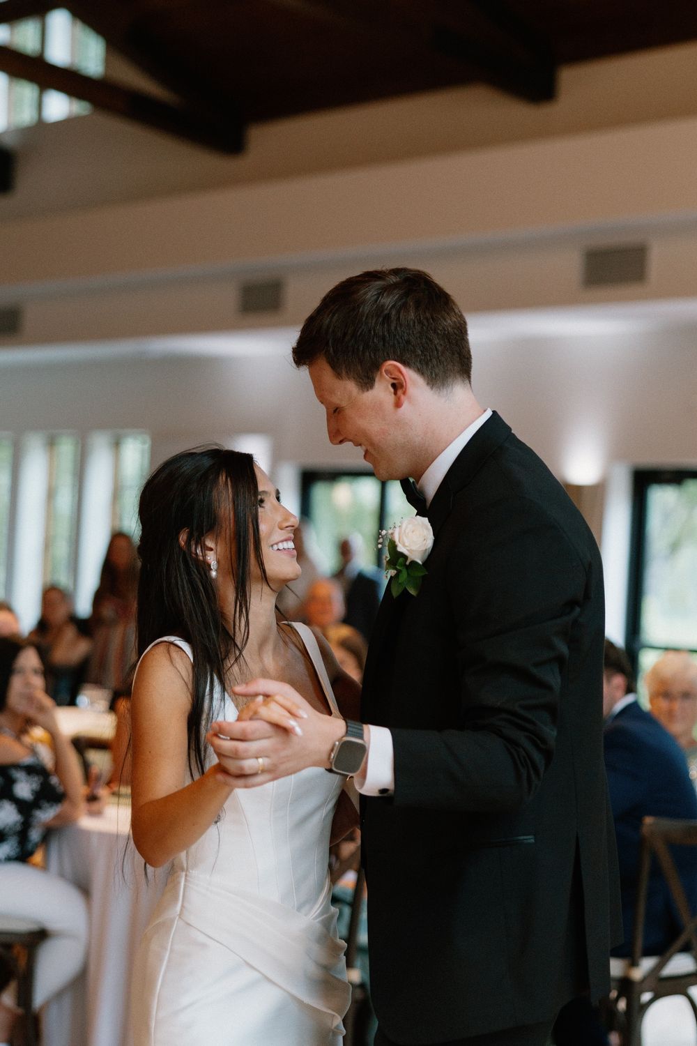 Bride and groom share their first dance, smiling joyfully, in an elegant wedding reception venue with guests seated nearby.
